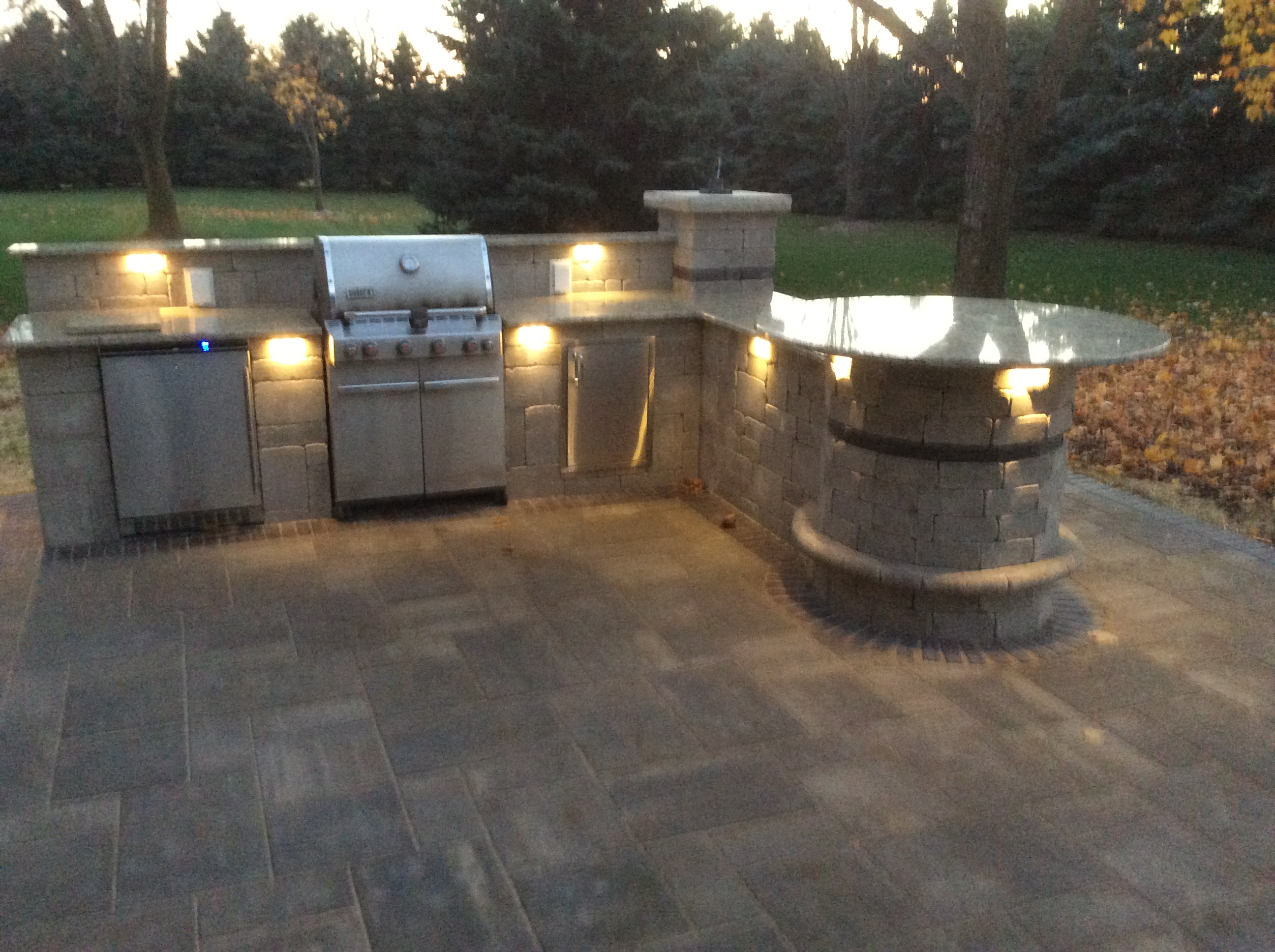 outdoor kitchen with grill and bar seating at dusk with lights on